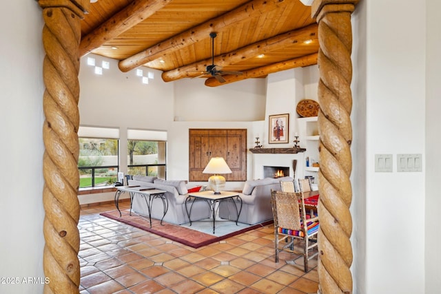 tiled living room featuring a lit fireplace, beam ceiling, wooden ceiling, a high ceiling, and a ceiling fan