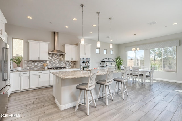 kitchen with wall chimney exhaust hood, hanging light fixtures, an island with sink, stainless steel appliances, and white cabinets