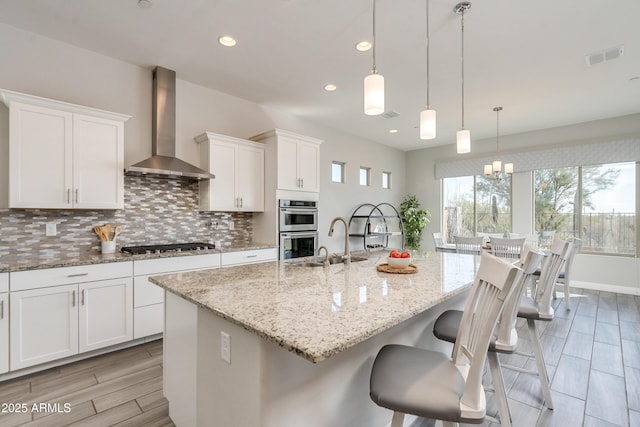 kitchen with white cabinetry, decorative light fixtures, a center island with sink, stainless steel appliances, and wall chimney range hood