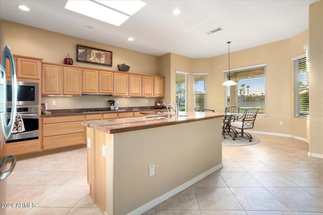 kitchen with stainless steel oven, hanging light fixtures, a kitchen island with sink, light brown cabinets, and light tile patterned flooring