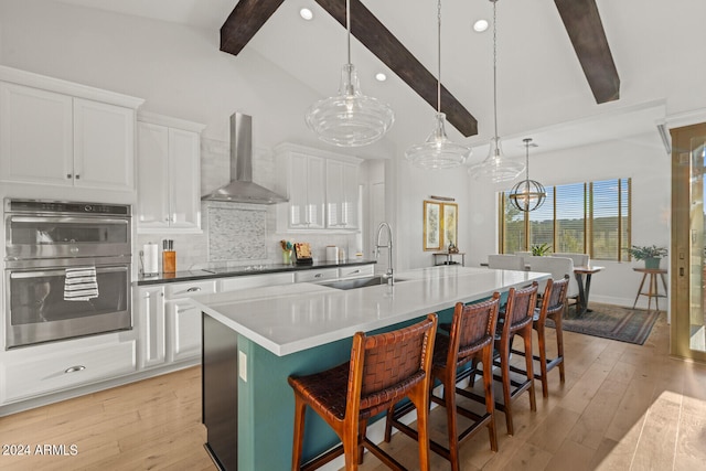 kitchen with double oven, beamed ceiling, wall chimney exhaust hood, and light wood-type flooring