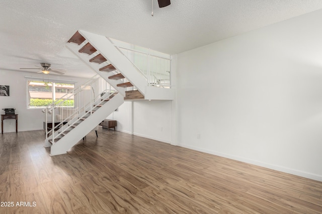 unfurnished living room with a ceiling fan, a textured ceiling, wood finished floors, stairway, and baseboards
