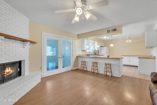 living area with a ceiling fan, a textured ceiling, french doors, light wood finished floors, and a brick fireplace