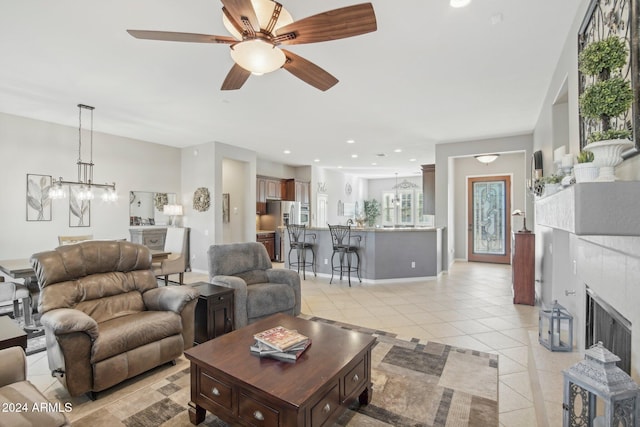 living room featuring a tiled fireplace, light tile patterned flooring, and ceiling fan with notable chandelier