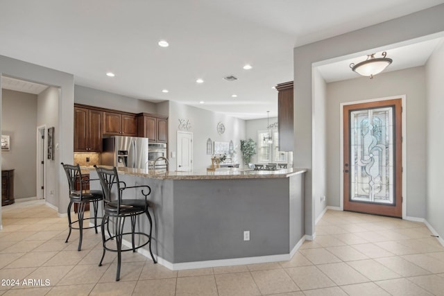 kitchen with light tile patterned flooring, kitchen peninsula, light stone countertops, a kitchen bar, and stainless steel fridge