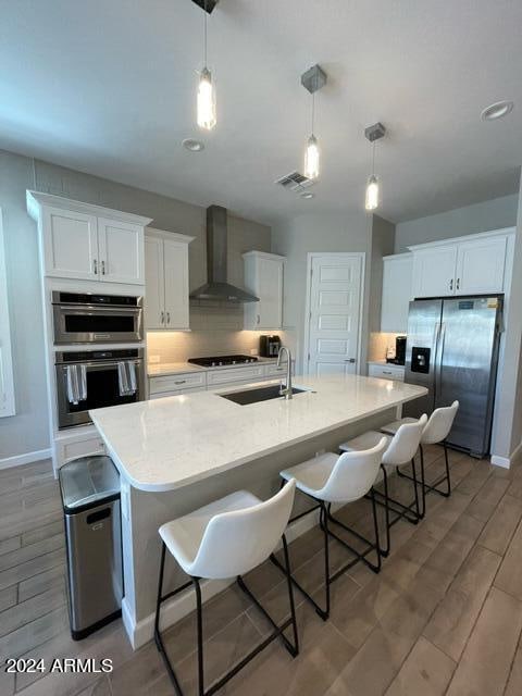 kitchen with an island with sink, appliances with stainless steel finishes, wall chimney exhaust hood, white cabinets, and sink