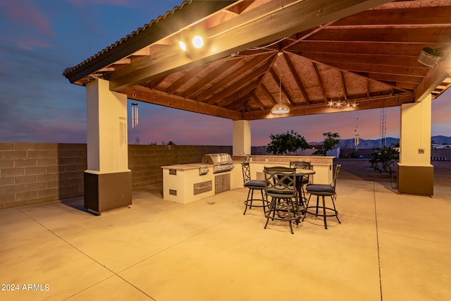 patio terrace at dusk with area for grilling, exterior bar, an outdoor kitchen, and a gazebo