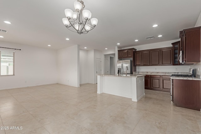 kitchen with appliances with stainless steel finishes, decorative light fixtures, an island with sink, light stone counters, and dark brown cabinets