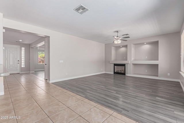 unfurnished living room with a glass covered fireplace, built in features, a ceiling fan, baseboards, and visible vents