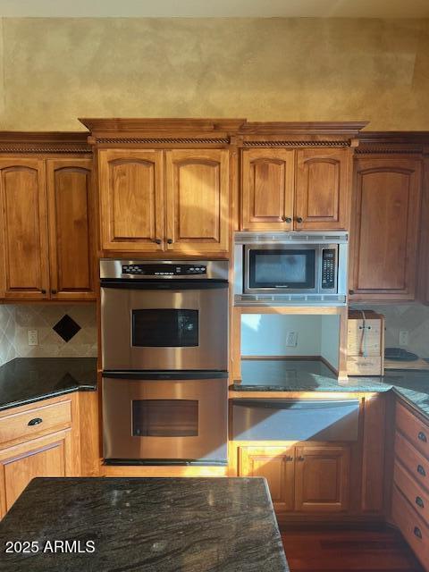 kitchen with backsplash, appliances with stainless steel finishes, brown cabinetry, and dark stone countertops