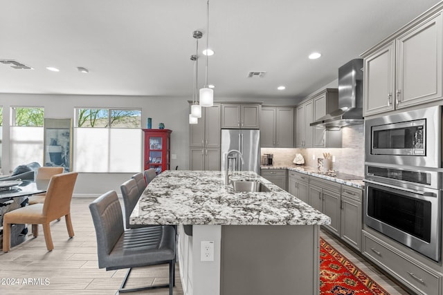 kitchen featuring gray cabinets, sink, wall chimney range hood, stainless steel appliances, and a center island with sink