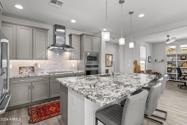 kitchen featuring light hardwood / wood-style floors, hanging light fixtures, wall chimney exhaust hood, stainless steel appliances, and ceiling fan