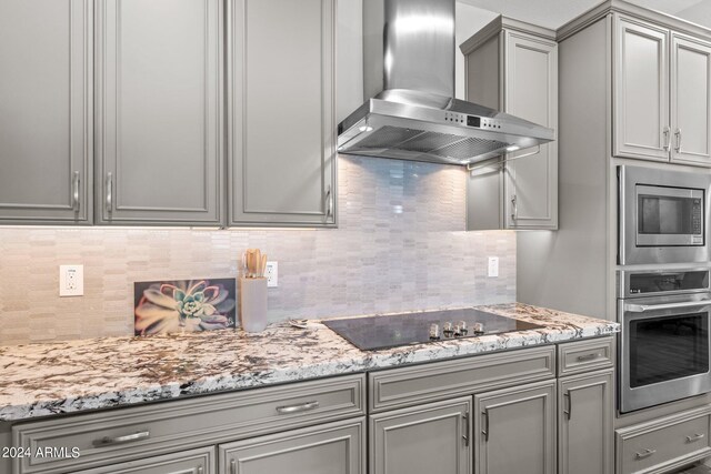 kitchen with gray cabinets, wall chimney exhaust hood, decorative backsplash, appliances with stainless steel finishes, and light stone countertops