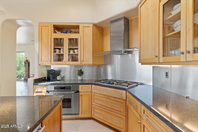 kitchen with dark stone countertops, stainless steel appliances, light brown cabinetry, decorative backsplash, and wall chimney exhaust hood