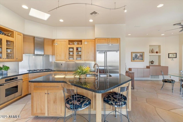 kitchen featuring appliances with stainless steel finishes, light brown cabinetry, an island with sink, a kitchen bar, and wall chimney exhaust hood