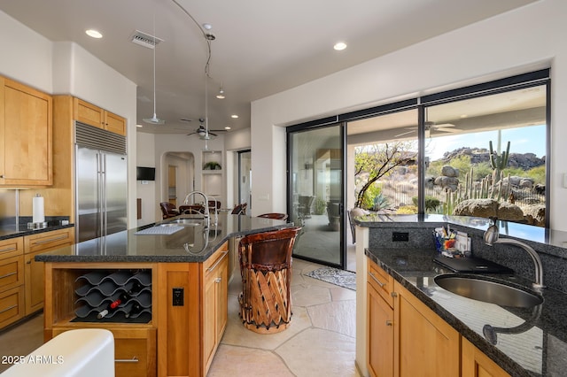 kitchen featuring stainless steel built in refrigerator, sink, dark stone counters, a kitchen island with sink, and ceiling fan