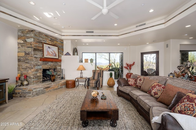 living room with a raised ceiling, a stone fireplace, and ceiling fan