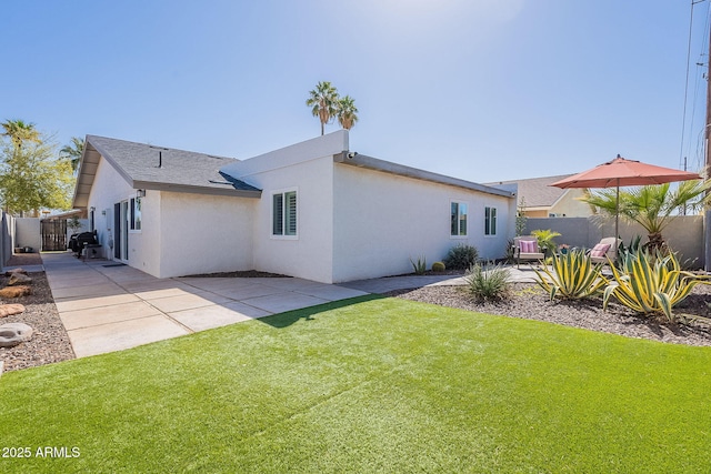 rear view of house featuring fence, roof with shingles, stucco siding, a lawn, and a patio area