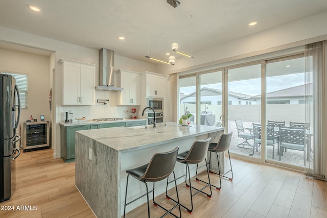 kitchen with a center island with sink, white cabinets, hanging light fixtures, wall chimney exhaust hood, and beverage cooler