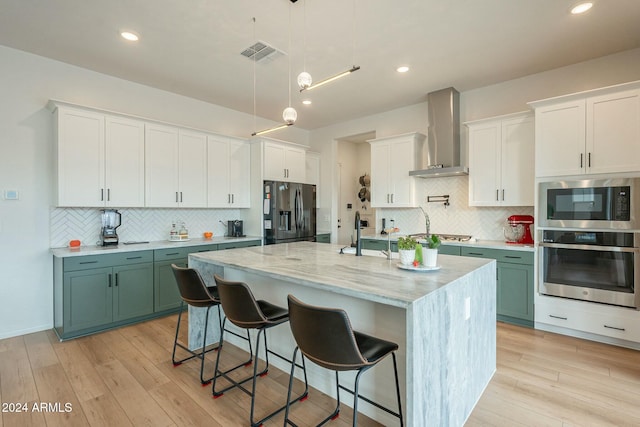 kitchen featuring light wood-type flooring, wall chimney range hood, an island with sink, and appliances with stainless steel finishes