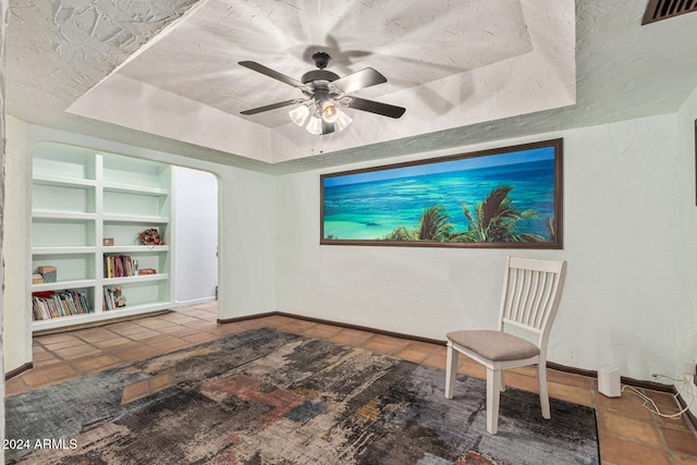 living area with ceiling fan, tile patterned flooring, built in features, a textured ceiling, and a tray ceiling