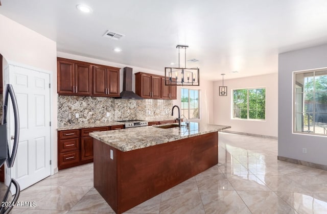 kitchen with pendant lighting, an island with sink, sink, wall chimney range hood, and light stone countertops