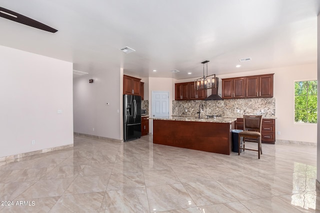 kitchen featuring an island with sink, hanging light fixtures, wall chimney range hood, light stone countertops, and black refrigerator with ice dispenser