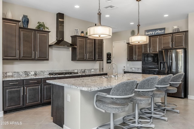 kitchen with wall chimney exhaust hood, dark brown cabinets, pendant lighting, a kitchen island with sink, and black appliances