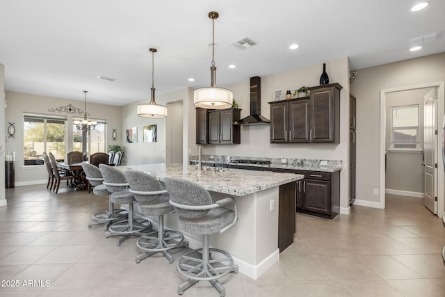 kitchen with a breakfast bar area, a kitchen island with sink, stainless steel gas cooktop, dark brown cabinetry, and wall chimney range hood