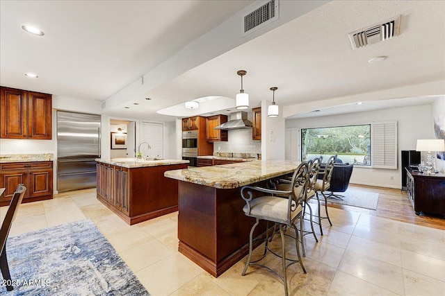 kitchen with wall chimney exhaust hood, sink, appliances with stainless steel finishes, an island with sink, and pendant lighting