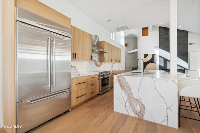 kitchen featuring high end appliances, a center island with sink, sink, wall chimney exhaust hood, and light hardwood / wood-style floors