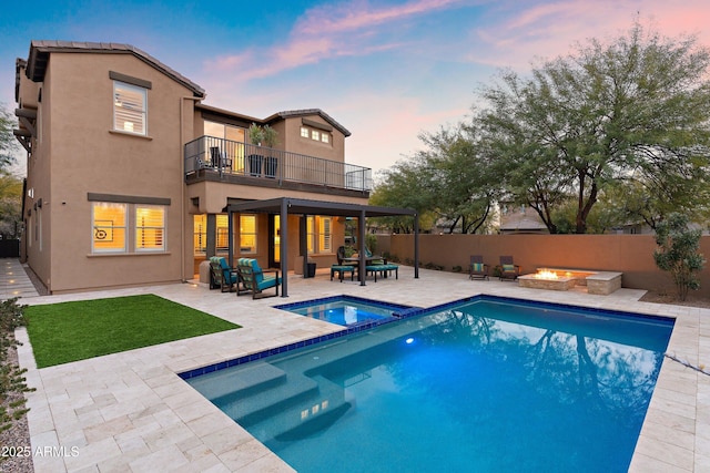 back house at dusk featuring a swimming pool with hot tub, a patio, a balcony, and an outdoor living space with a fire pit