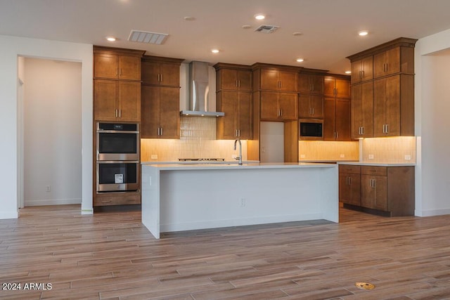 kitchen featuring a kitchen island with sink, light hardwood / wood-style flooring, wall chimney range hood, and appliances with stainless steel finishes