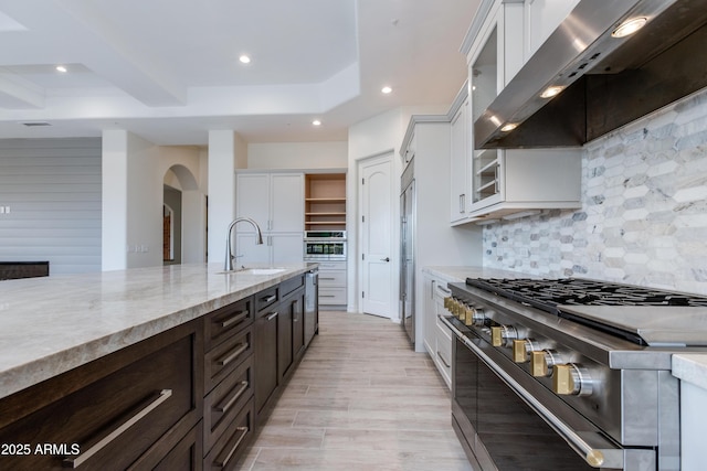 kitchen with premium appliances, a sink, white cabinetry, wall chimney range hood, and glass insert cabinets