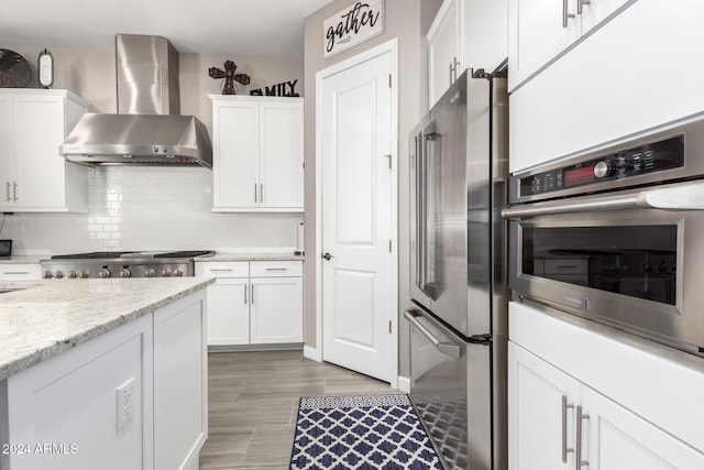 kitchen with stainless steel appliances, light stone countertops, hardwood / wood-style flooring, white cabinets, and wall chimney exhaust hood