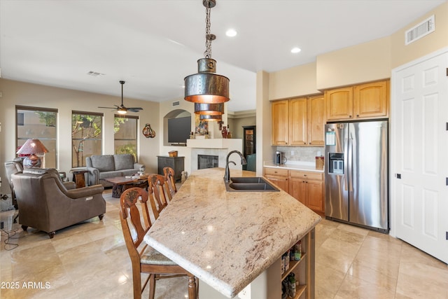 kitchen featuring a center island with sink, sink, stainless steel refrigerator with ice dispenser, ceiling fan, and a tiled fireplace