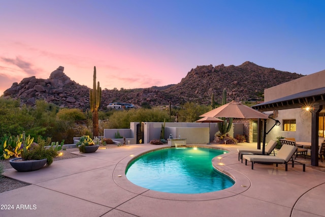 pool at dusk featuring a mountain view, pool water feature, and a patio