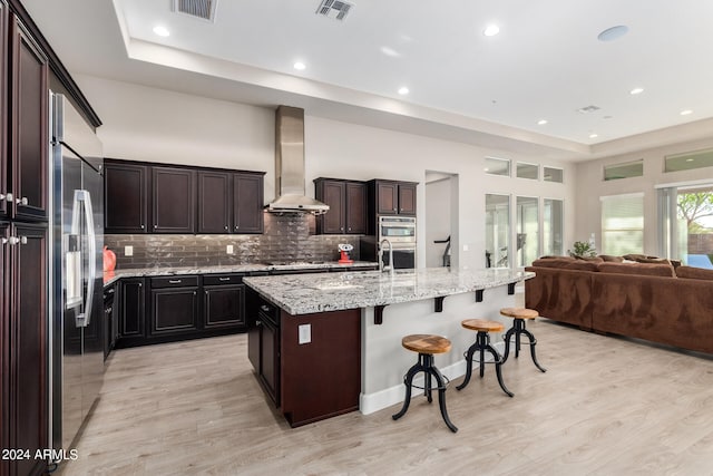 kitchen featuring light hardwood / wood-style floors, a kitchen island with sink, wall chimney range hood, tasteful backsplash, and a breakfast bar