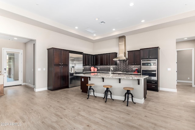 kitchen with wall chimney exhaust hood, light hardwood / wood-style flooring, a kitchen island with sink, a breakfast bar, and stainless steel appliances