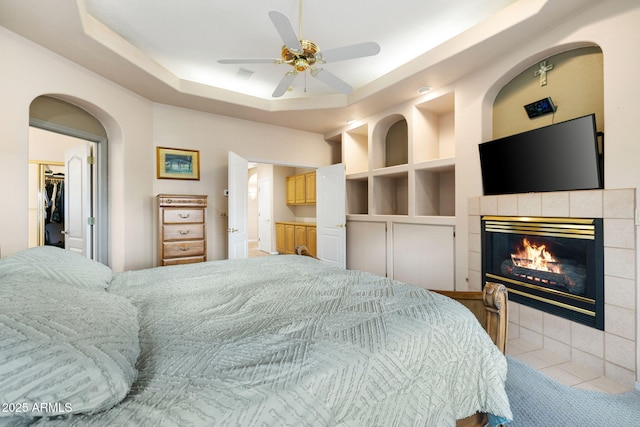 bedroom featuring ensuite bathroom, a tiled fireplace, a tray ceiling, ceiling fan, and a spacious closet