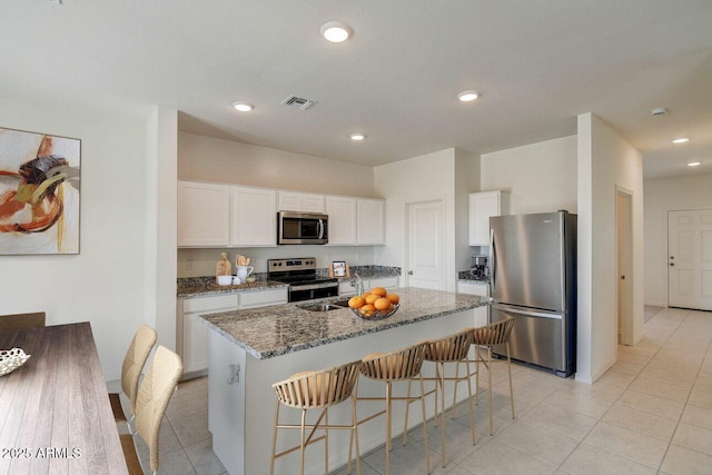 kitchen with dark stone counters, a kitchen island with sink, stainless steel appliances, a kitchen bar, and white cabinetry