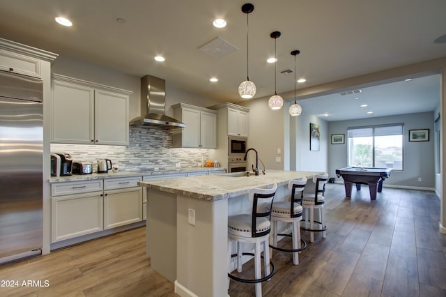 kitchen with stainless steel appliances, decorative light fixtures, an island with sink, wall chimney range hood, and billiards