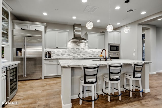 kitchen featuring stainless steel appliances, an island with sink, hanging light fixtures, beverage cooler, and wall chimney range hood