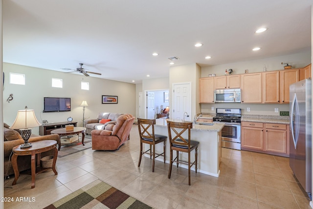 kitchen with stainless steel appliances, a kitchen breakfast bar, light stone counters, an island with sink, and light brown cabinetry