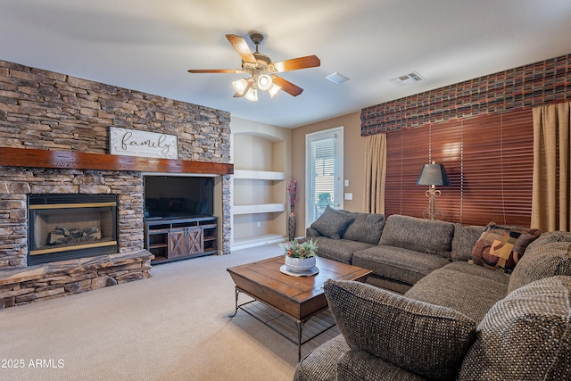 living room featuring built in shelves, ceiling fan, a stone fireplace, and light colored carpet