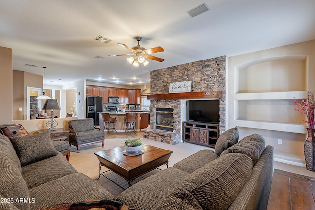 living room with built in features, light colored carpet, a stone fireplace, and ceiling fan