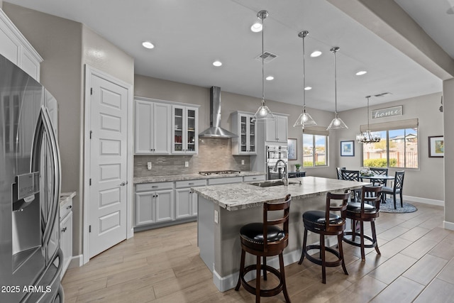 kitchen with appliances with stainless steel finishes, white cabinets, hanging light fixtures, a kitchen island with sink, and wall chimney range hood