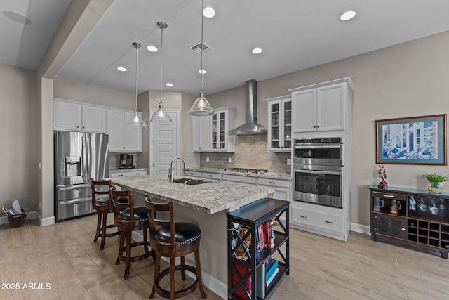 kitchen with appliances with stainless steel finishes, an island with sink, white cabinets, hanging light fixtures, and wall chimney range hood