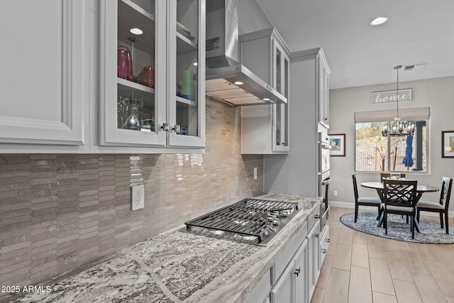 kitchen featuring white cabinetry, appliances with stainless steel finishes, a notable chandelier, light stone countertops, and wall chimney range hood