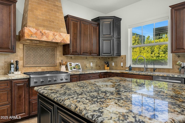 kitchen featuring a sink, appliances with stainless steel finishes, dark stone counters, wall chimney exhaust hood, and tasteful backsplash